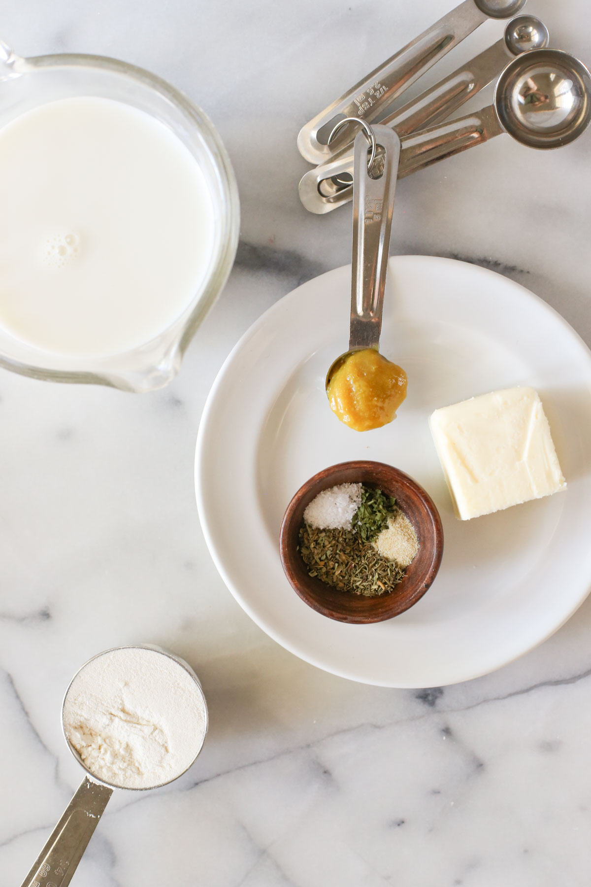 Overhead shot of the ingredients for Homemade Cream of Chicken Soup - a glass measuring cup of milk and flour in a measuring cup both sitting next to a plate with butter, a teaspoon full of the chicken base and a little wood bowl with the spices in it.