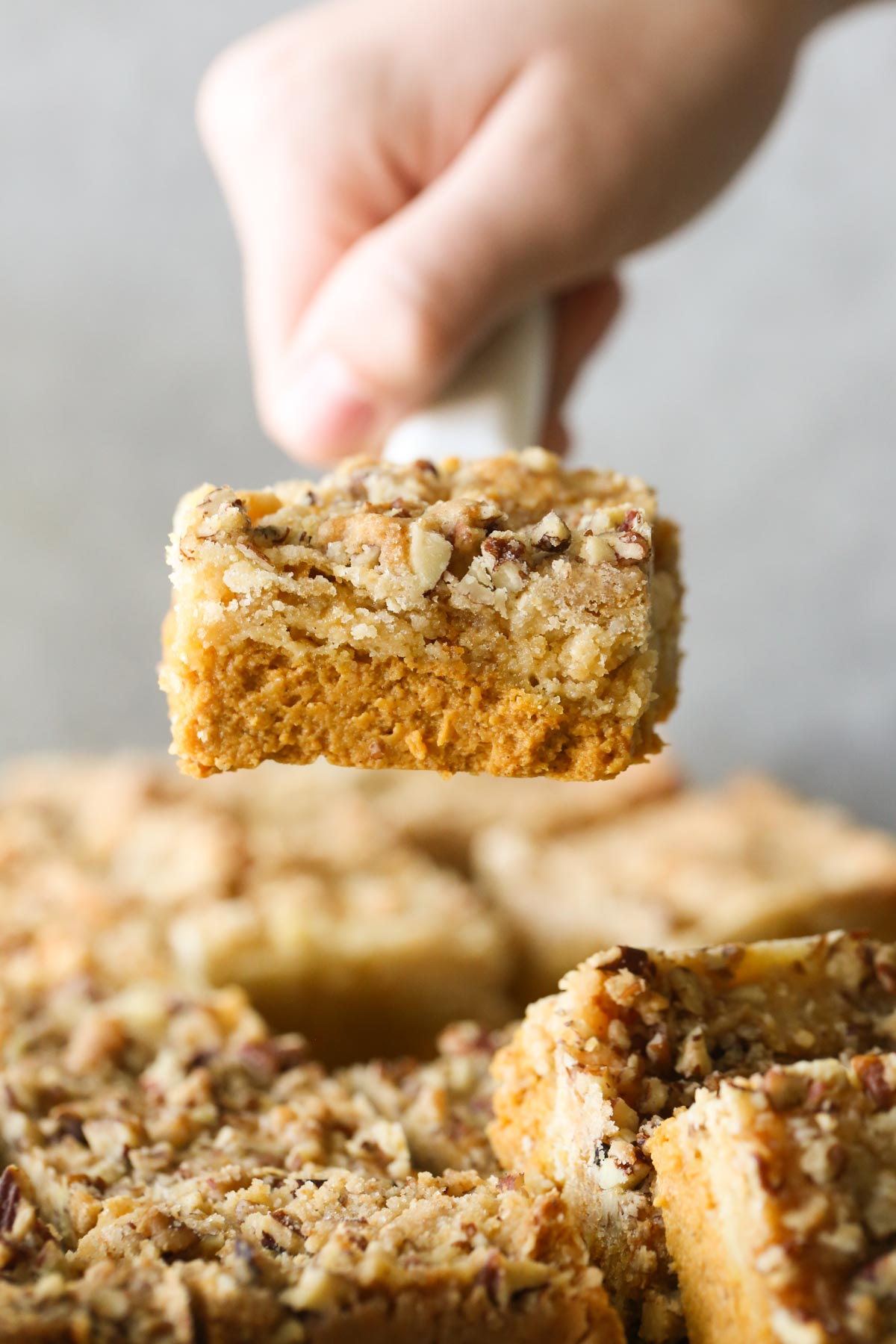 Close up shot of a hand holding a spatula with a Pumpkin Pie Bar on it above the rest of the cut Pumpkin Pie Bars. 