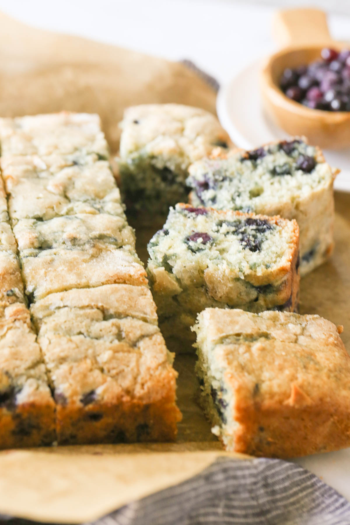 Close up shot of Sourdough Blueberry Breakfast Cake on parchment paper that has been sliced into pieces, with a wood scoop of wild blueberries in the background. 