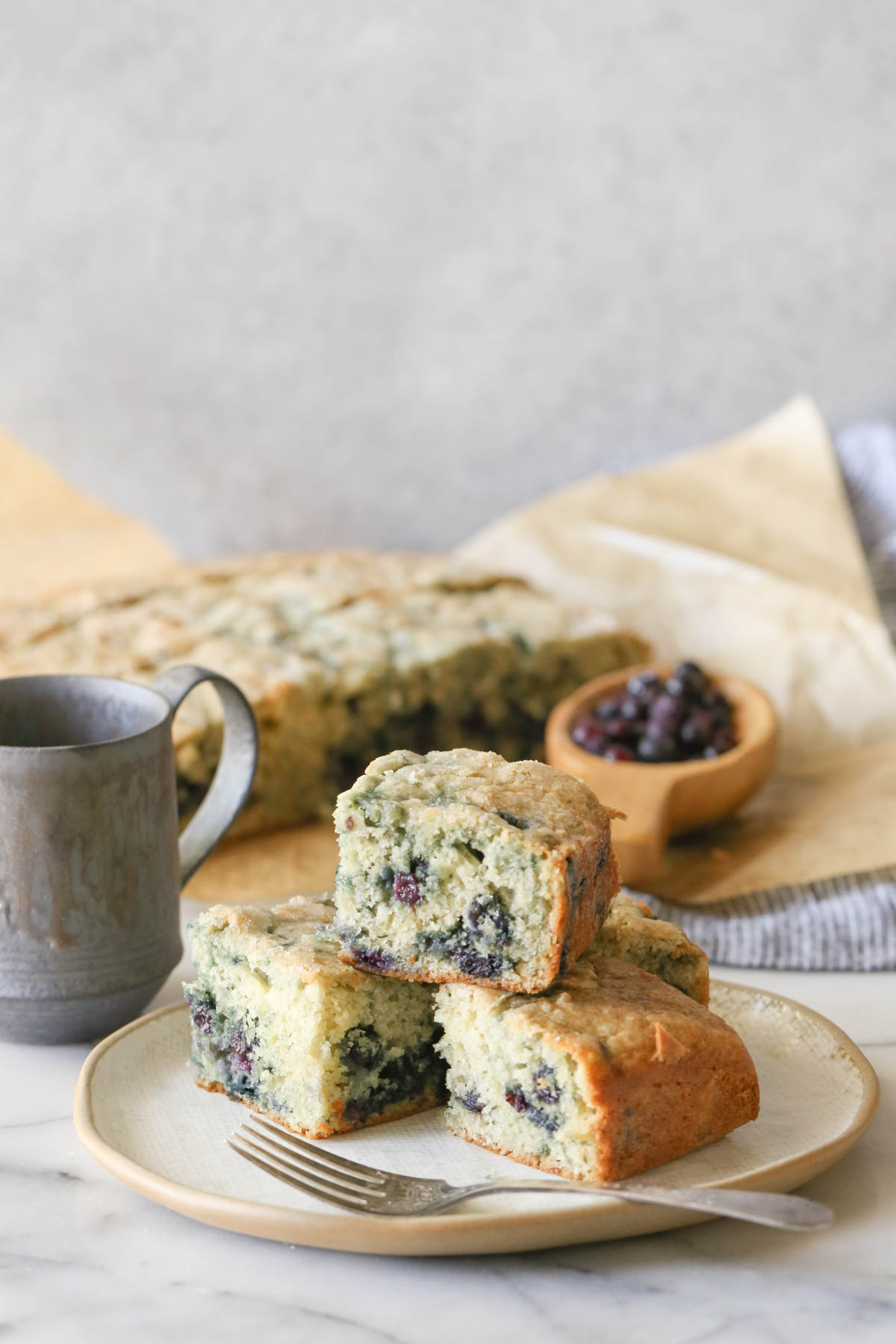 Four pieces of Sourdough Blueberry Breakfast Cake on a plate with a fork, with a coffee mug and the rest of the Sourdough Blueberry Breakfast Cake in the background, along with a wood scoop of wild blueberries. 