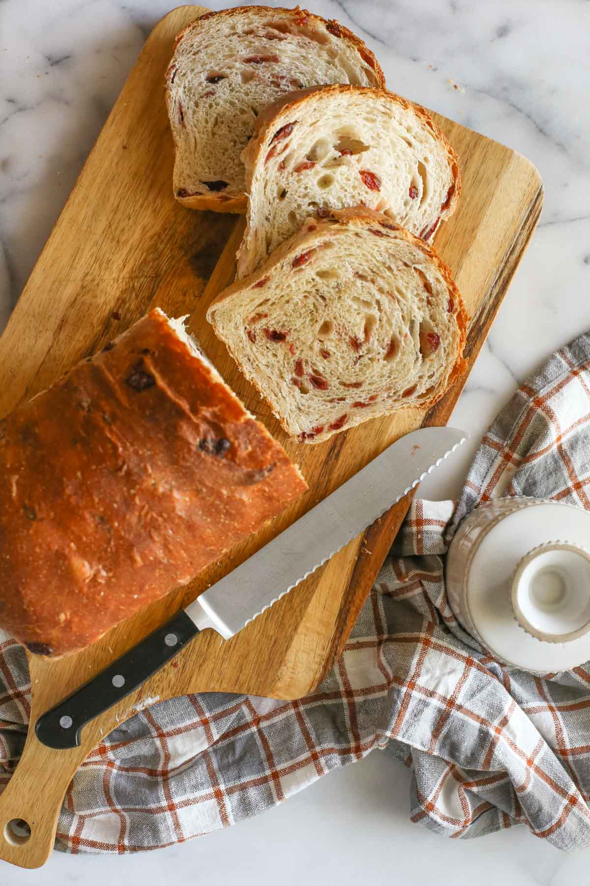 Overhead shot of a loaf of Sourdough Cranberry Bread on a cutting board with a serrated knife and three slice of bread cut from the loaf, and a butter crock next the the cutting board, all sitting on a plaid dish towel.