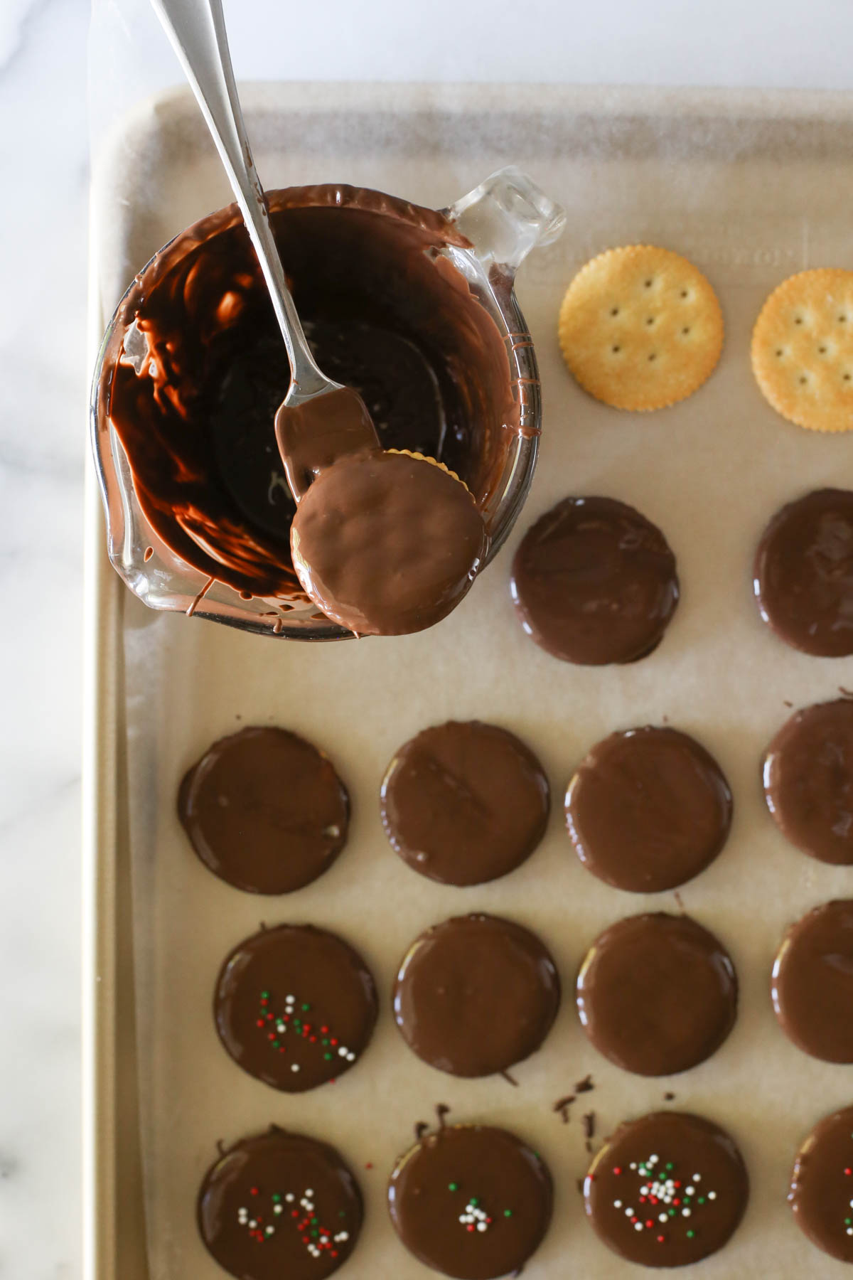 Overhead shot of a Ritz cracker on a fork that has just been dipped in a glass measuring cup of the melted chocolate mixture, with more Copycat Thin Mints on wax paper, some with sprinkles and some without, and two Ritz crackers that have not been coated yet. 