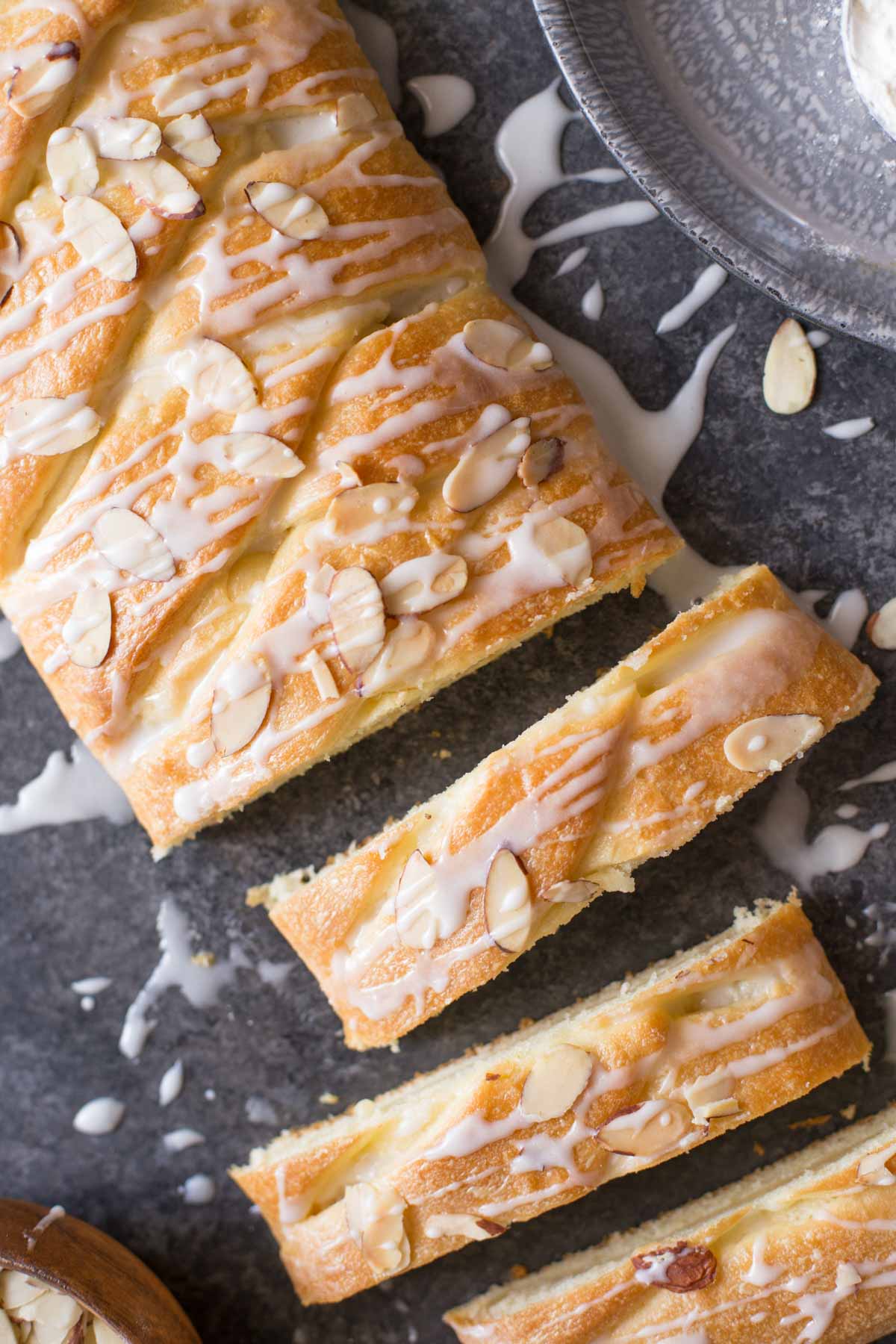 Close up overhead shot of the Almond Braid with three slices cut away from it and a small wood bowl of sliced almonds next to the slices, and a metal tray with flour next to the Almond Braid. 