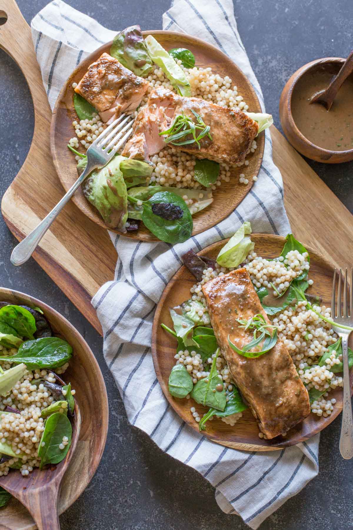 Overhead shot of two wood plates with a fillet of Honey Mustard Balsamic Roasted Salmon on a bed of Couscous Salad, with a wood bowl of the Couscous Salad next to the plates, along with a small wood bowl of the Honey Mustard Balsamic marinade.  