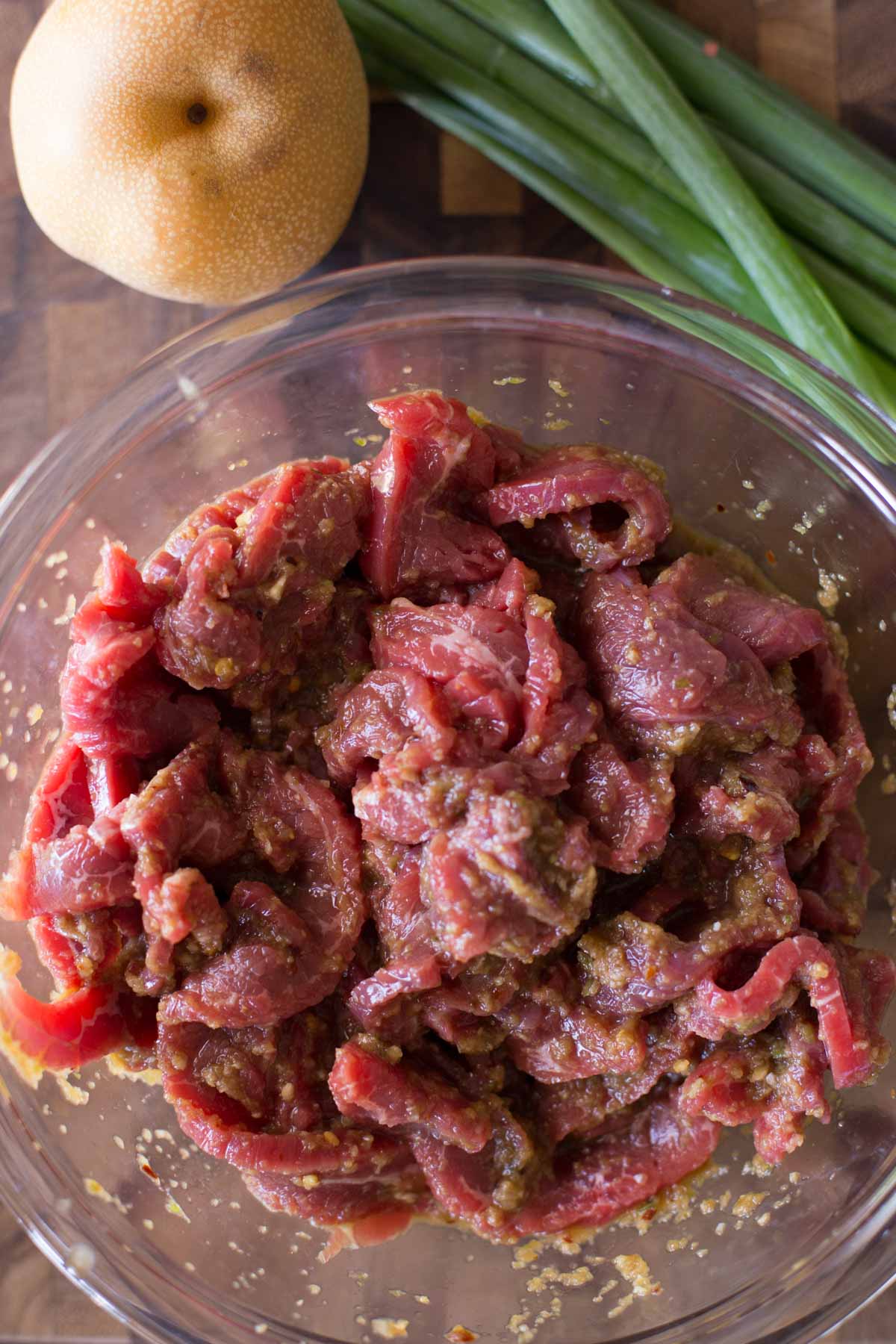 Overhead shot of the skirt steak marinating in a glass bowl, with an Asian pear and some green onions next to the bowl.
