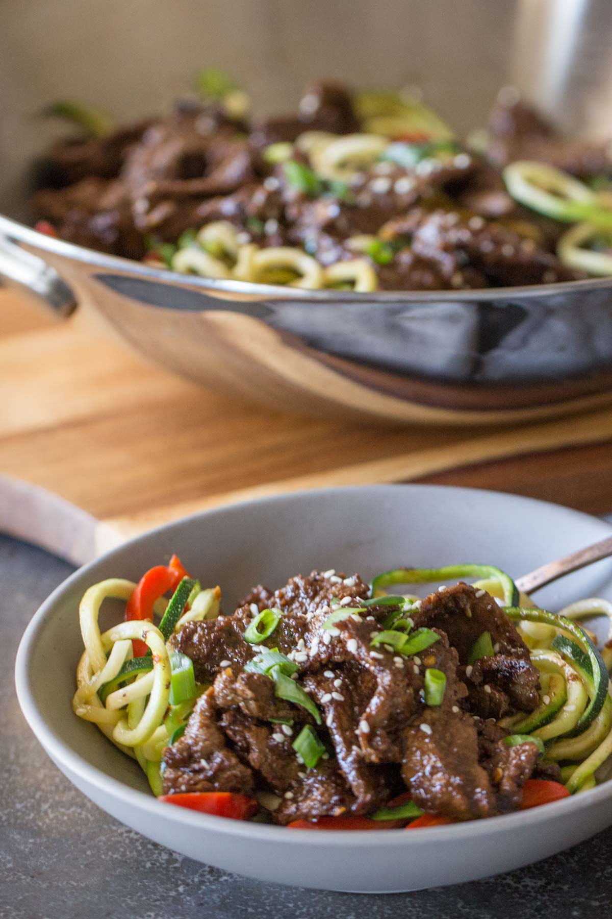 Close up shot of a bowl of Spicy Korean Beef and Zoodles, with a large wok of Spicy Korean Beef and Zoodles in the background.