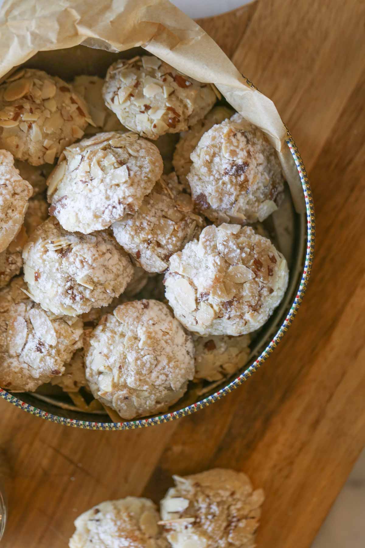 Overhead shot of Almond Paste Cookies in a parchment paper lined cookie tin, sitting on a wood cutting board, with more cookies on the board. 