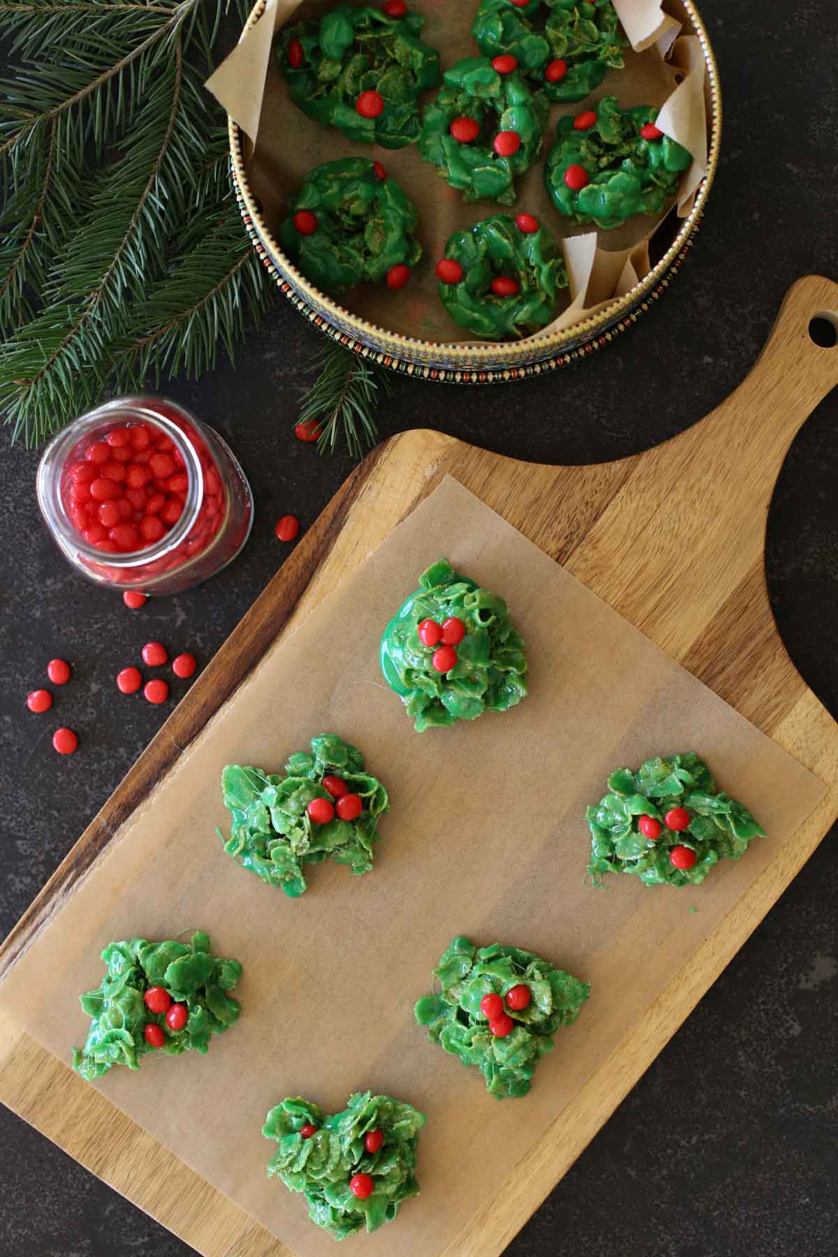 Overhead shot of six Holly Cookies on parchment paper on a wood cutting board, with more Holly Cookies in a cookie tin lined with parchment paper next to the cutting board, along with a small glass jar of cinnamon candies. 