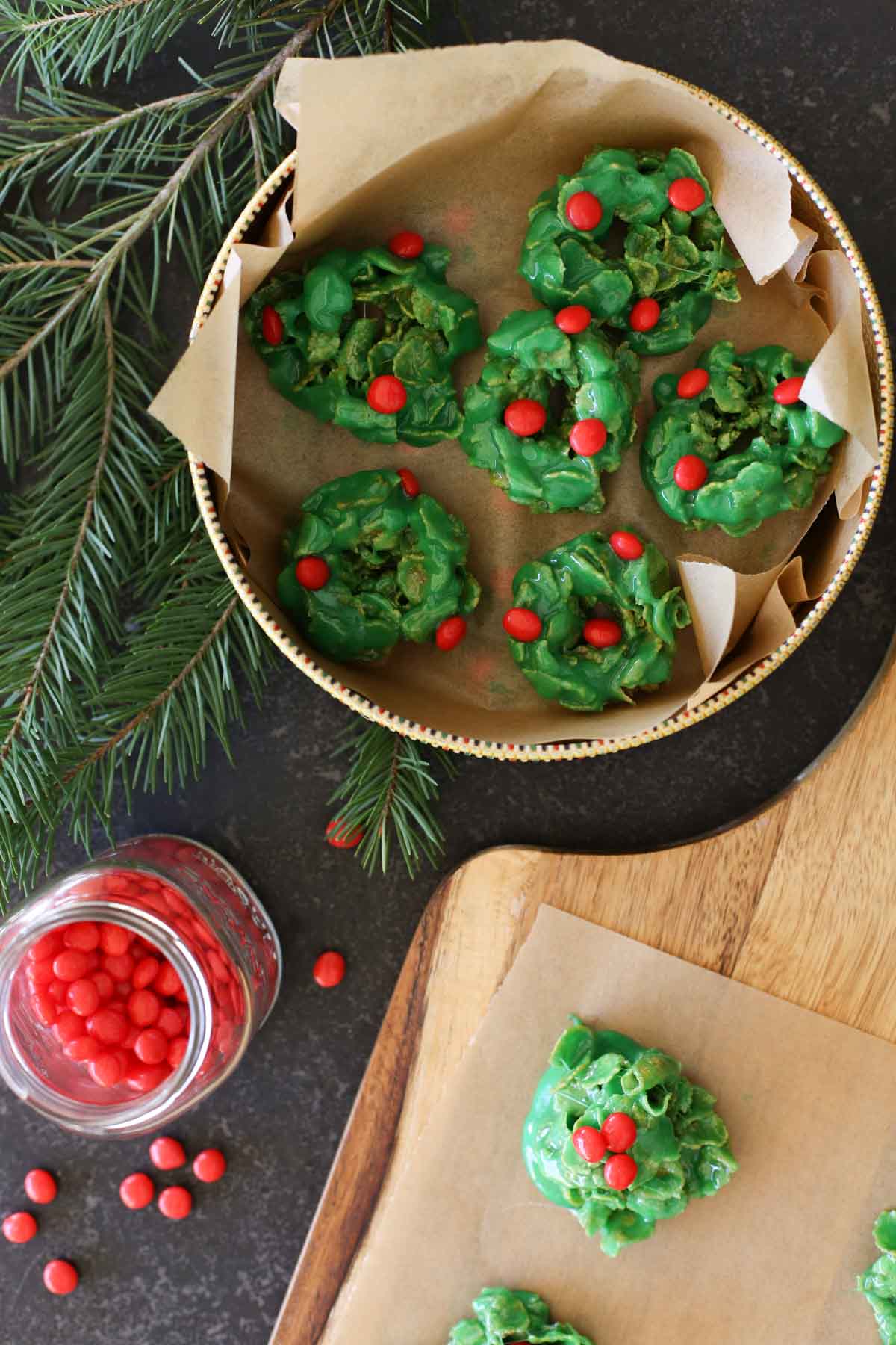 Overhead shot of Holly Cookies in a cookie tin lined with parchment paper, next to a wood cutting bard with more Holly Cookies on parchment paper, and a small glass jar of cinnamon candies.  