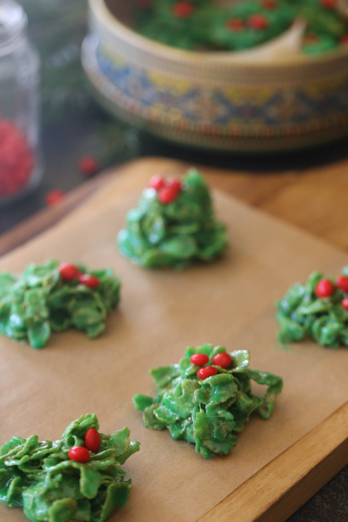 Holly Cookies on parchment paper on a wood cutting board, with a cookie tin of Holly Cookies in the background. 