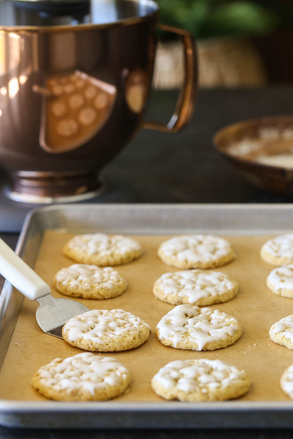 Iced Lemon Oatmeal Cookies on a parchment paper lined baking sheet, with a spatula under one cookie, and a mixing bowl in the background.