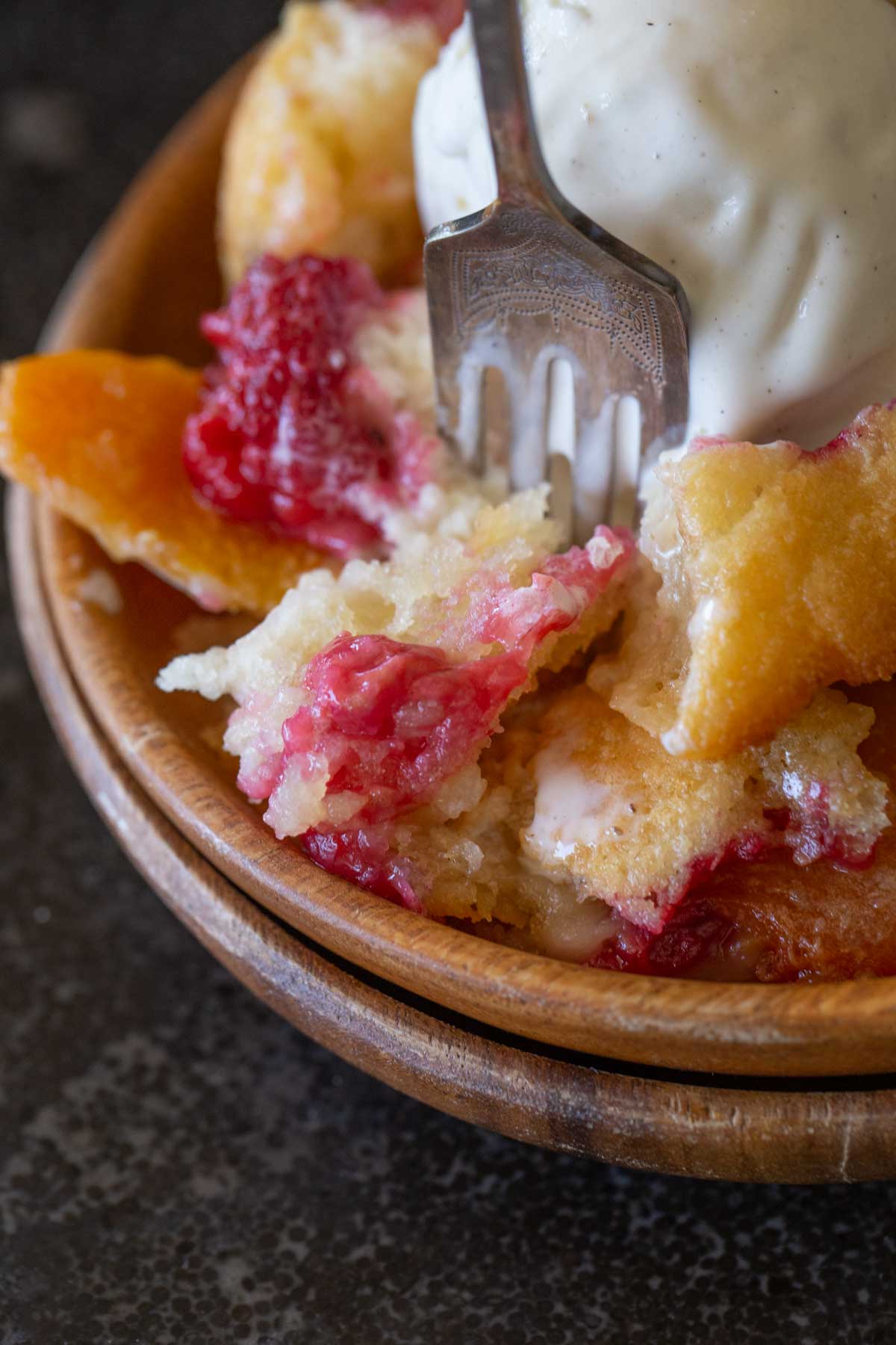 Close up shot of a fork in a serving of Sourdough Peach Cobbler topped with vanilla ice cream in a wood bowl.