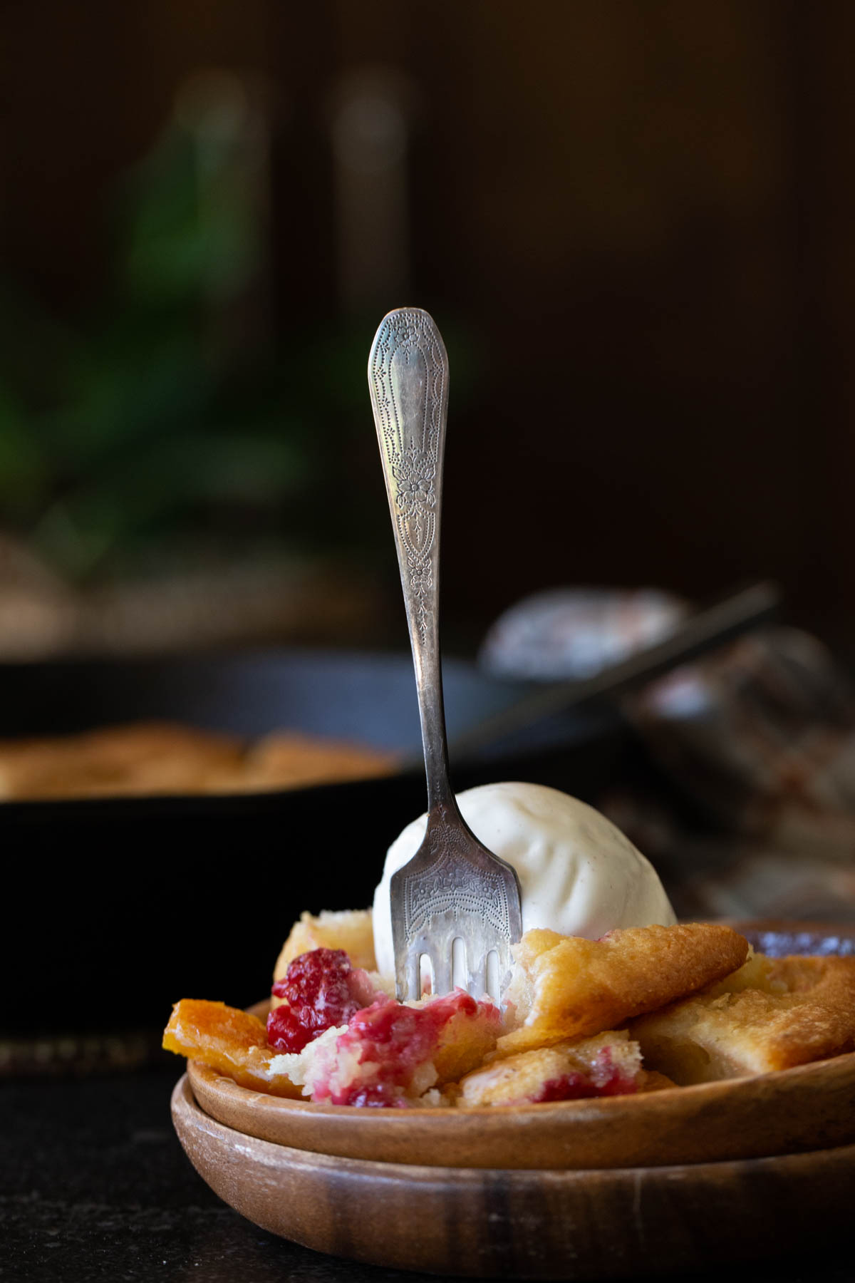 A fork stuck into a serving of Sourdough Peach Cobbler topped with vanilla ice cream in a wood bowl.