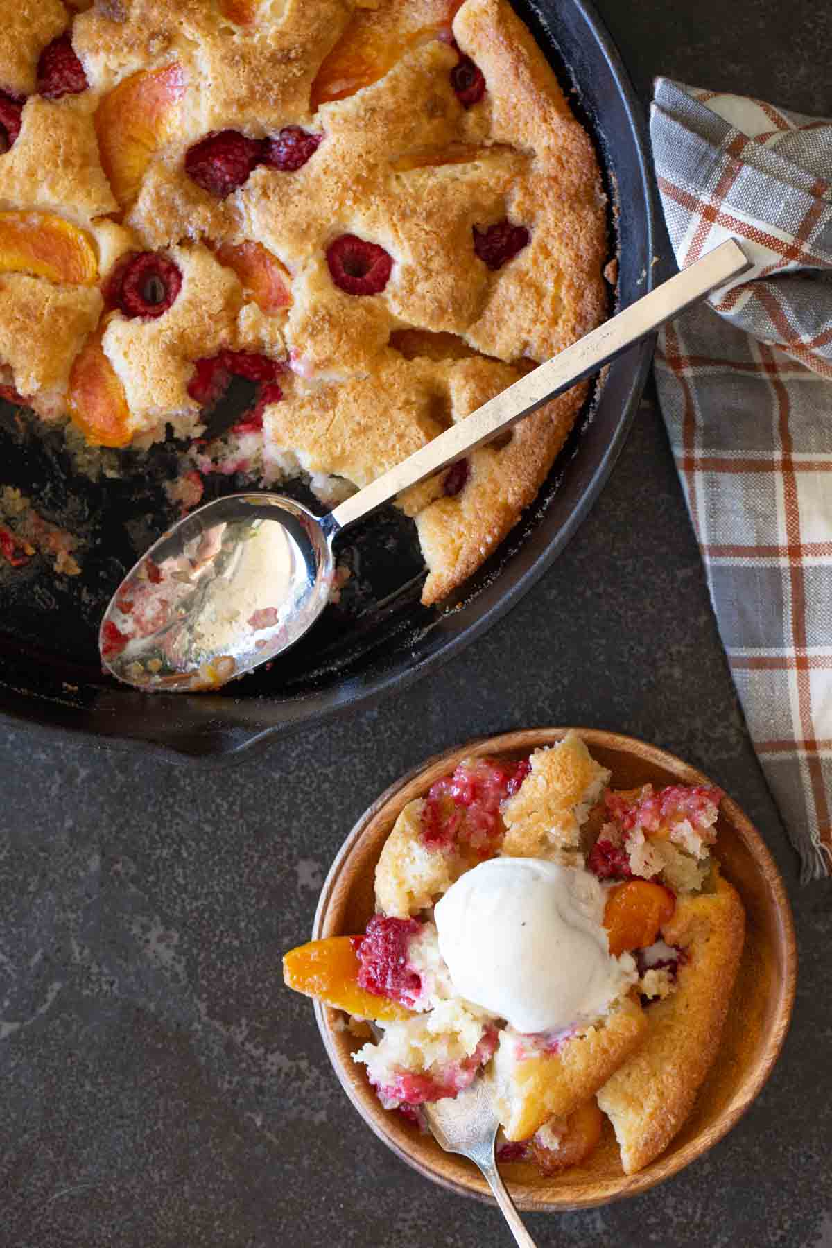 Overhead shot of Sourdough Peach Cobbler in a cast iron skillet, with a serving of Sourdough Peach Cobbler topped with vanilla ice cream in a wood bowl next to the skillet.