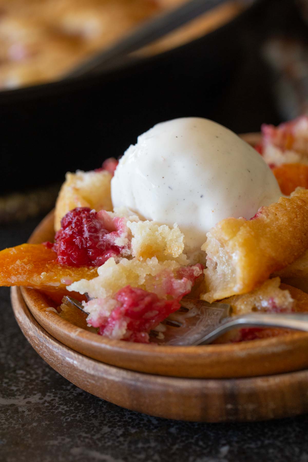 Close up shot of a serving of Sourdough Peach Cobbler topped with vanilla ice cream in a wood bowl.