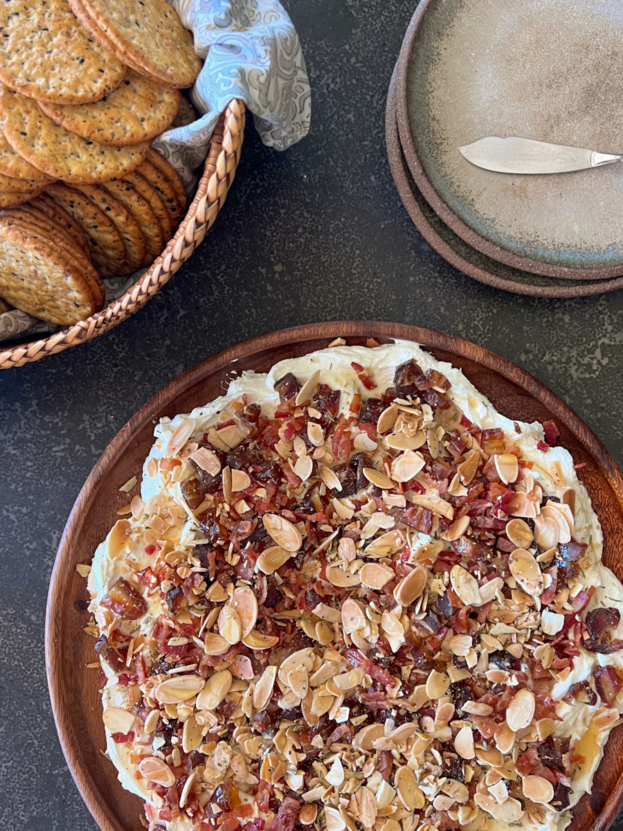 Overhead shot of Whipped Goat Cheese with Bacon, Dates and Almonds on a wooden serving plate, with a basket of crackers and a stack of plates with a spreading knife next to it.