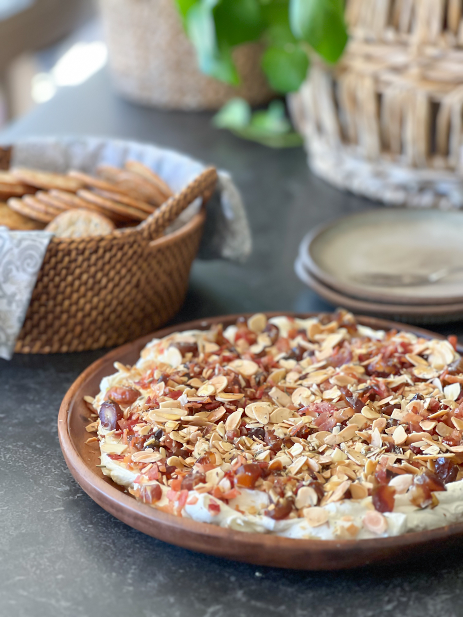 Whipped Goat Cheese with Bacon, Dates and Almonds on a wood serving plate, with a basket of crackers and a stack of plates in the background.