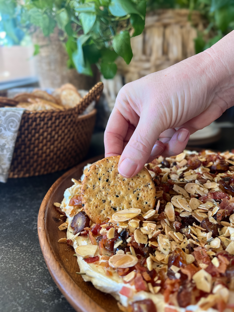 A hand dipping a cracker in the Whipped Goat Cheese with Bacon, Dates and Almonds, with a basket of crackers in the background.