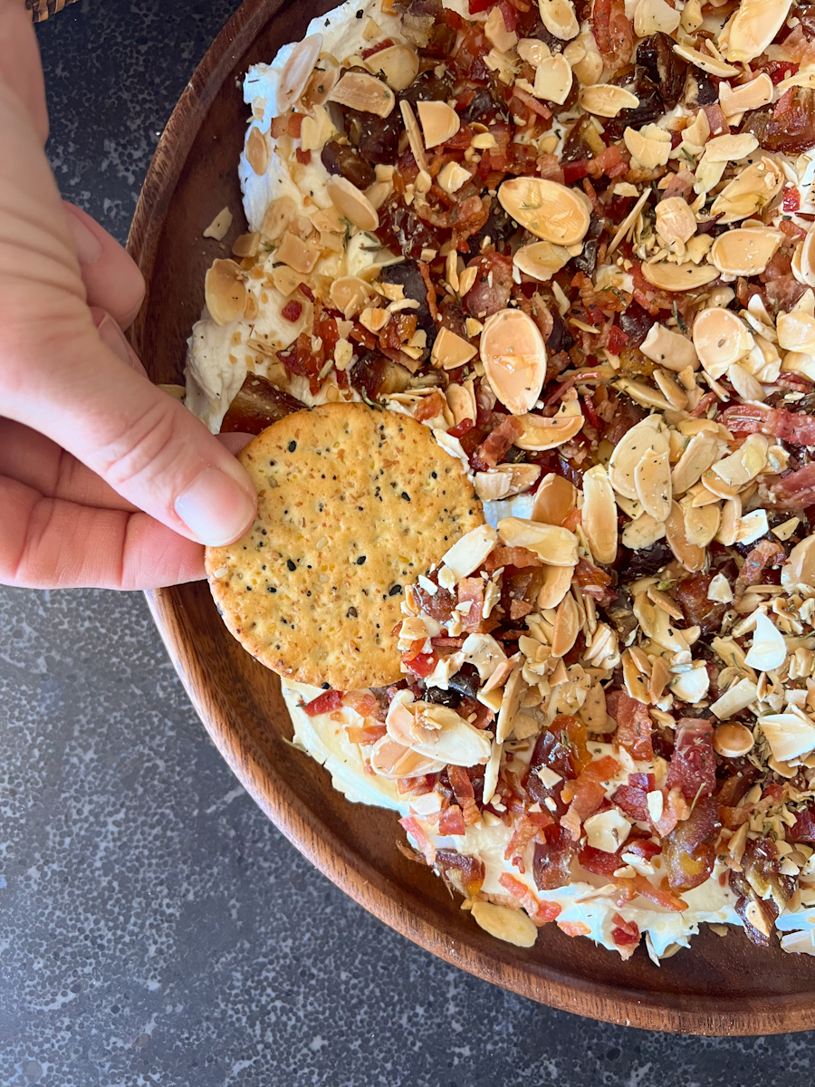 Overhead closeup shot of a hand dipping a cracker into the Whipped Goat Cheese with Bacon, Dates and Almonds.
