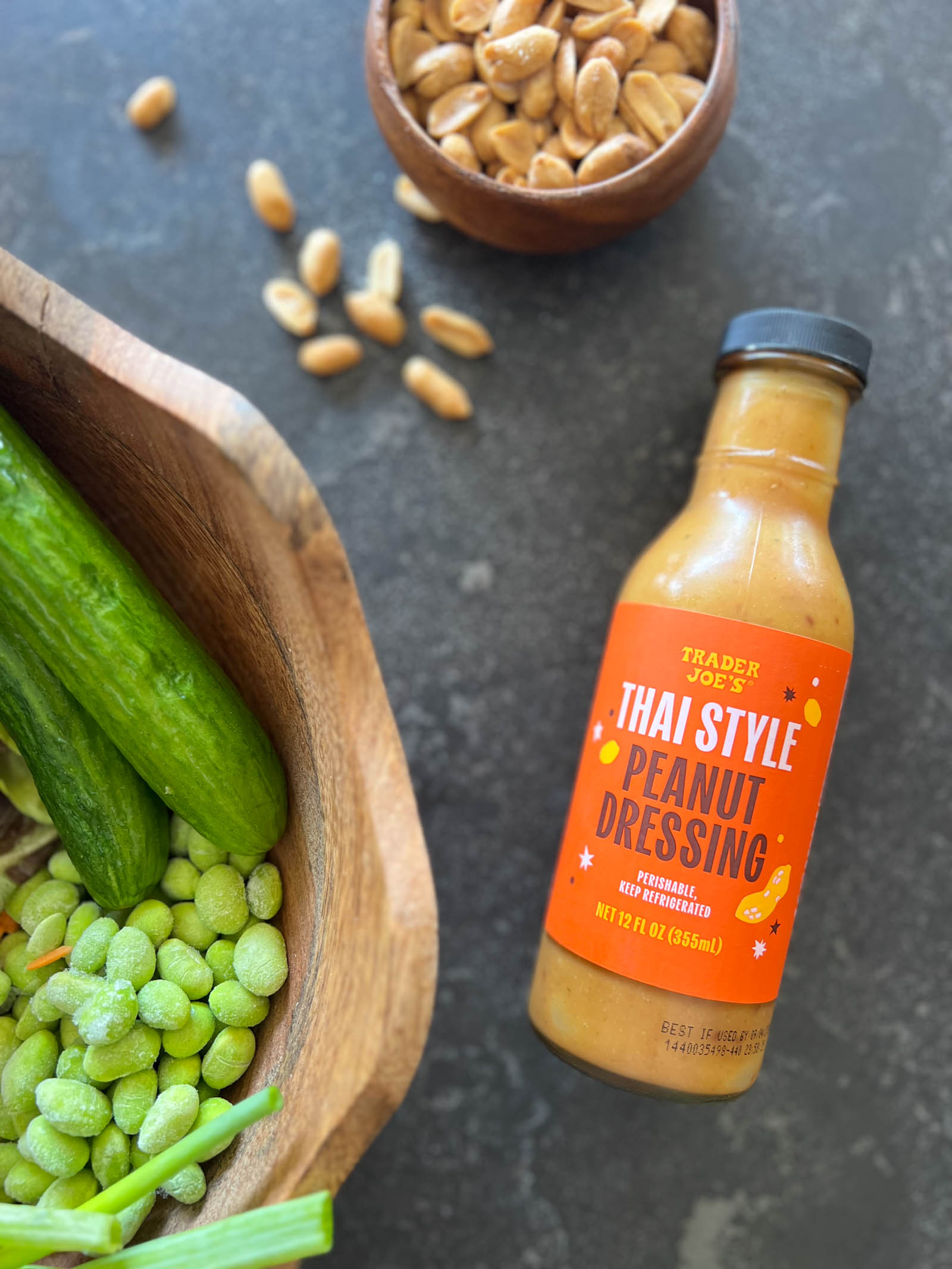 Overhead shot of a bottle of Trader Joe's Thai Style Peanut Dressing laying next to a small wood bowl of peanuts and a large wood bowl of cucumbers, edamame, and green onions.  