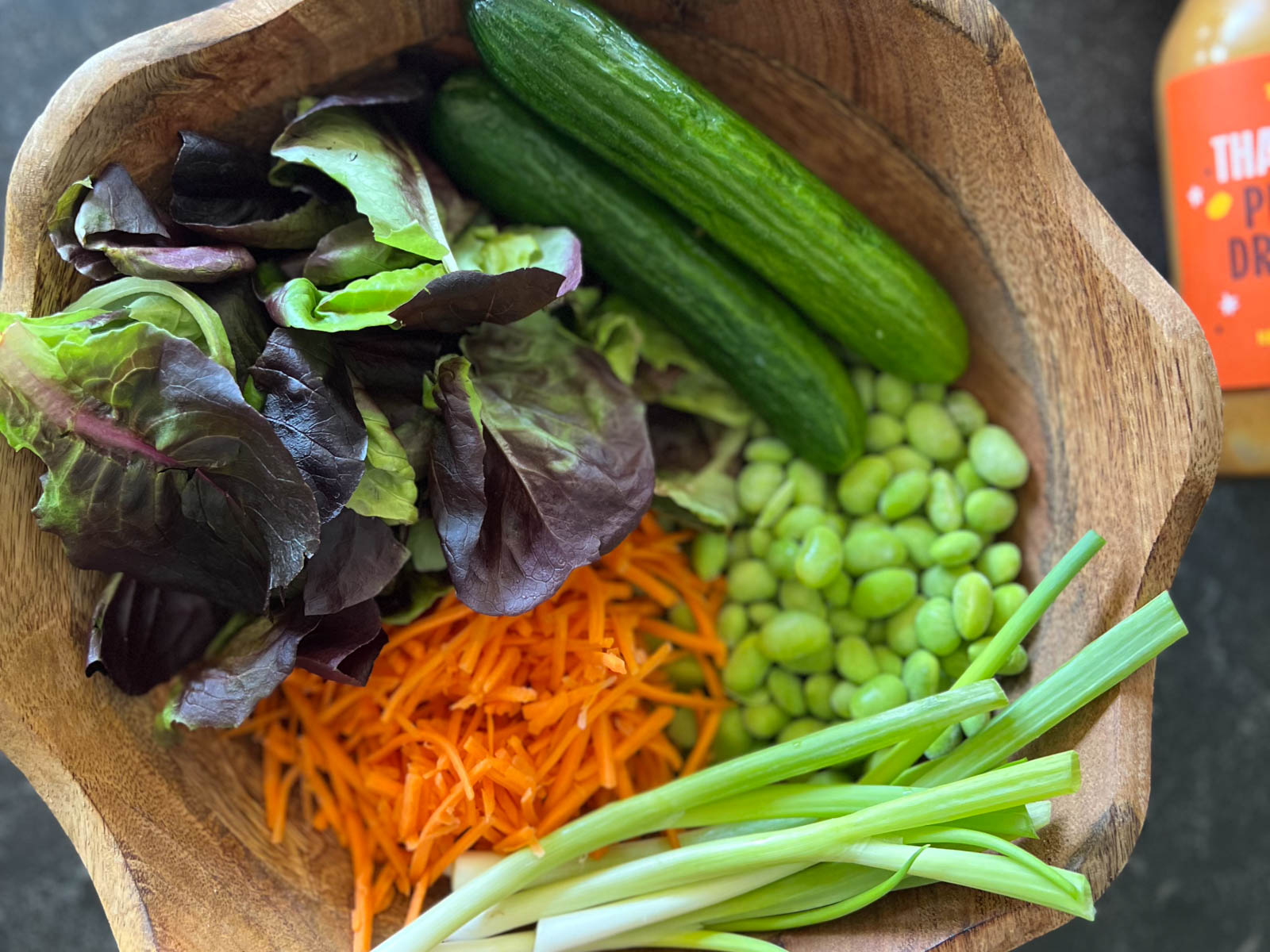 Overhead shot of a wood bowl with butter lettuce, cucumbers, edamame, shredded carrots, and green onions, which a jar of dressing next to it.  