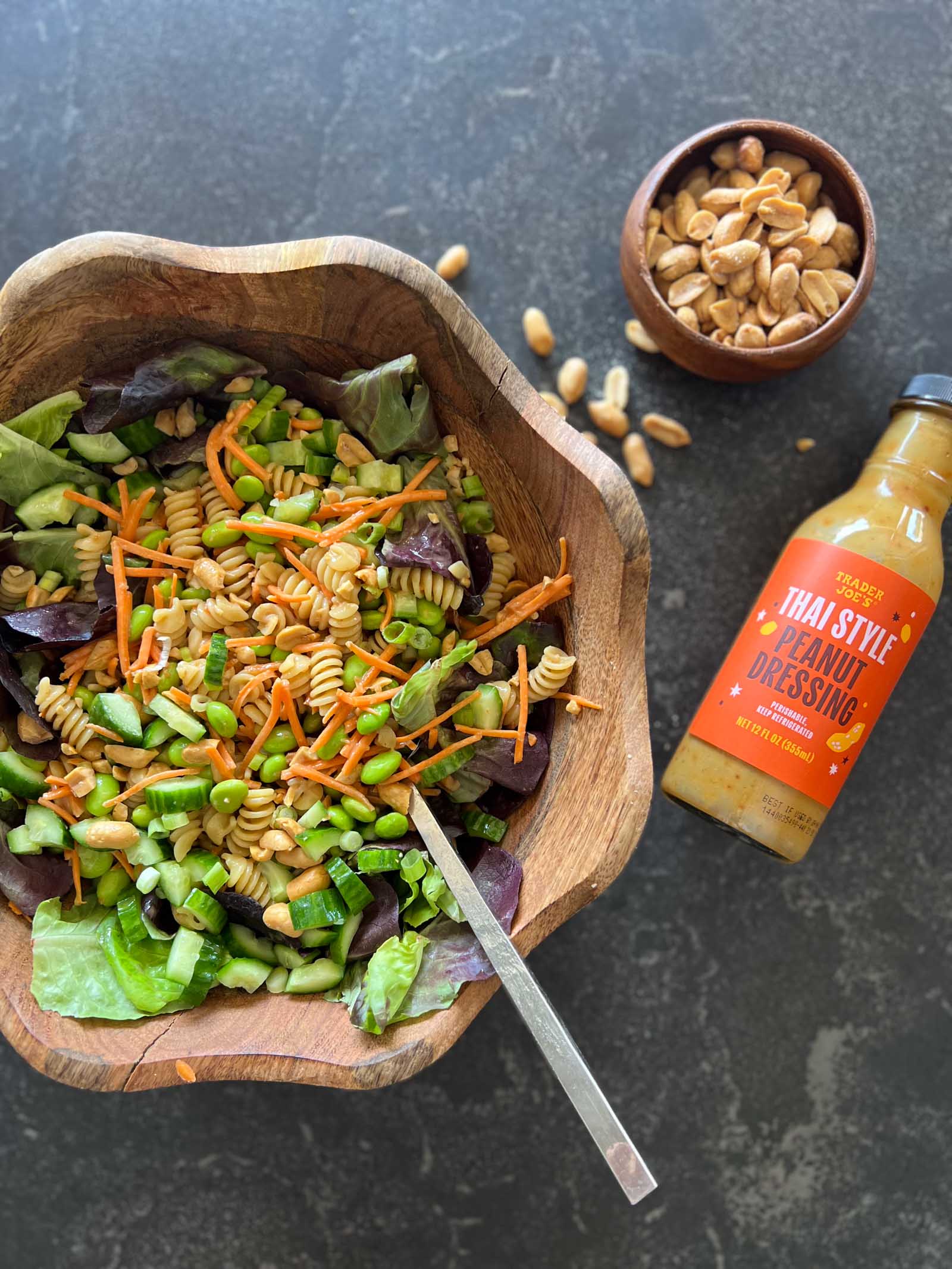Overhead shot of Thai Peanut Pasta Salad in a wood bowl with of a bottle of Trader Joe's Thai Style Peanut Dressing laying next to it along with a small wood bowl of peanuts.