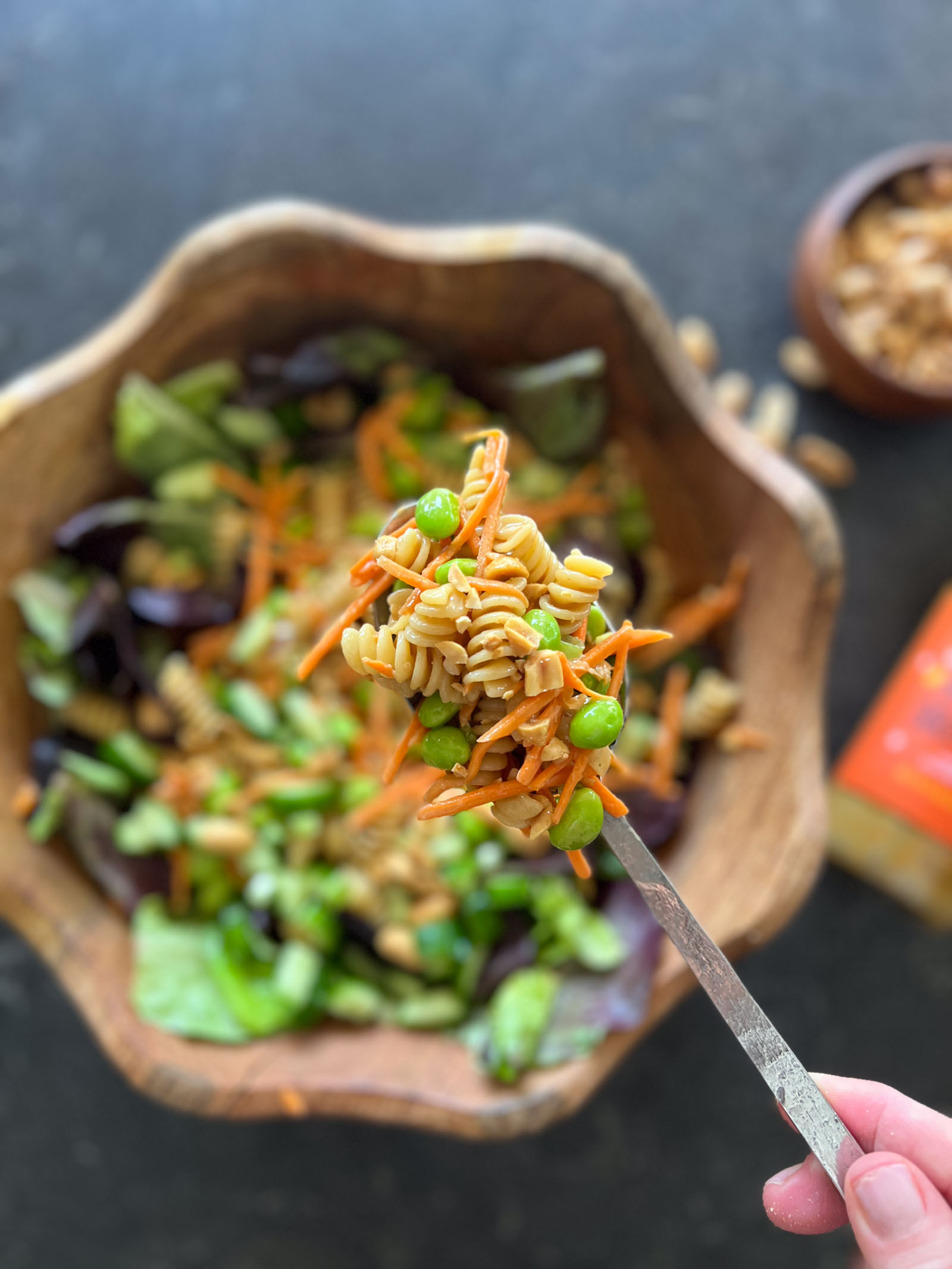 Close up shot of a spoonful of Thai Peanut Pasta Salad, with a wood bowl  of Thai Peanut Pasta Salad in the background, and a bottle of Trader Joe's Thai Style Peanut Dressing laying next to it along with a small wood bowl of peanuts.