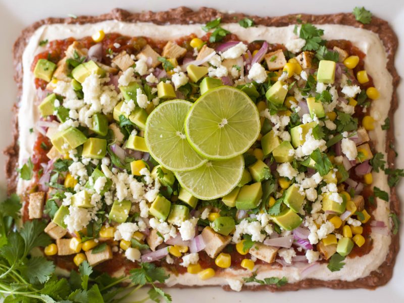 Overhead view of nine layer dip on a white rectangular platter, topped with diced avocado, corn, red onion, cilantro, crumbled queso fresco, and lime slices.