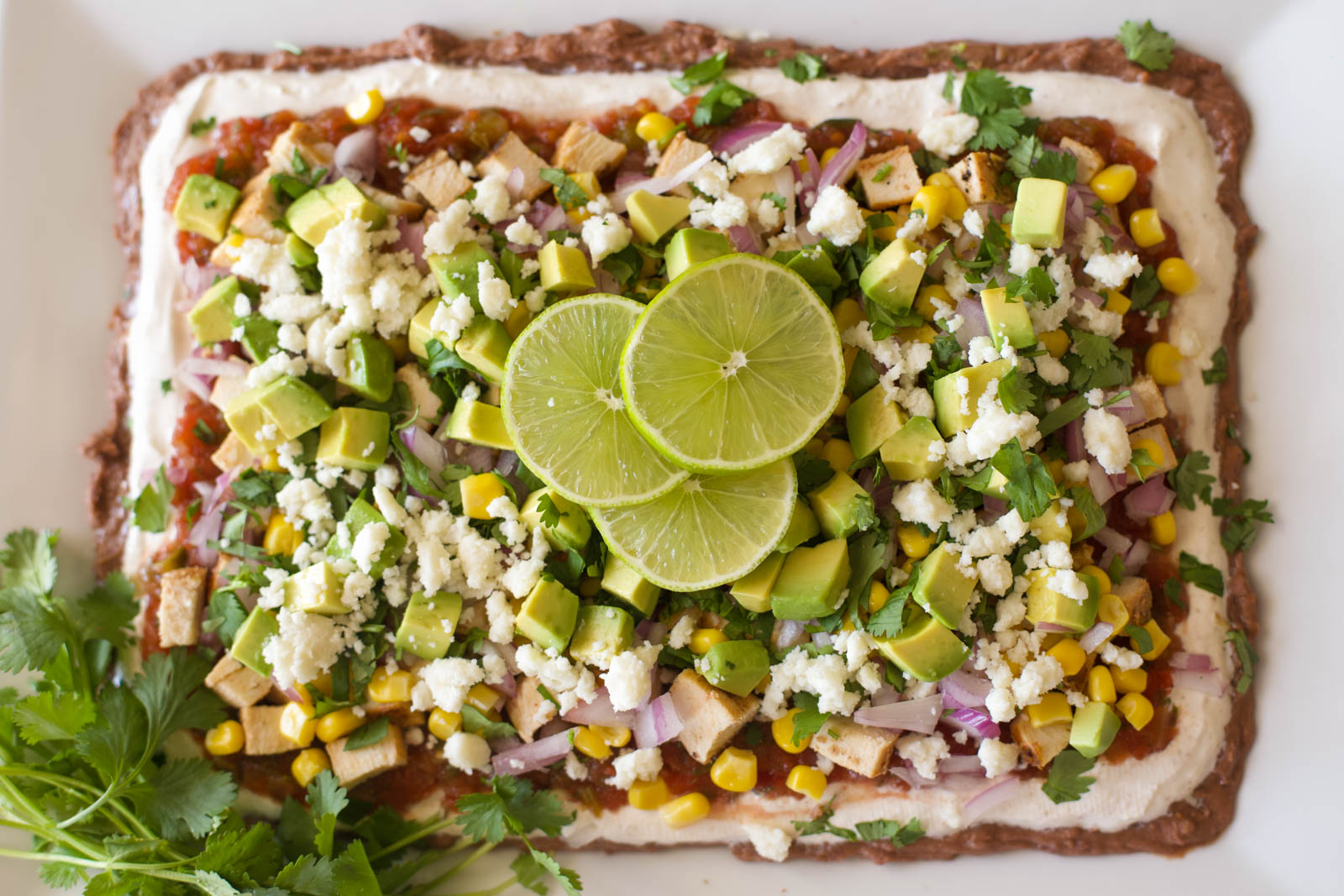 Overhead view of nine layer dip on a white rectangular platter, topped with diced avocado, corn, red onion, cilantro, crumbled queso fresco, and lime slices.