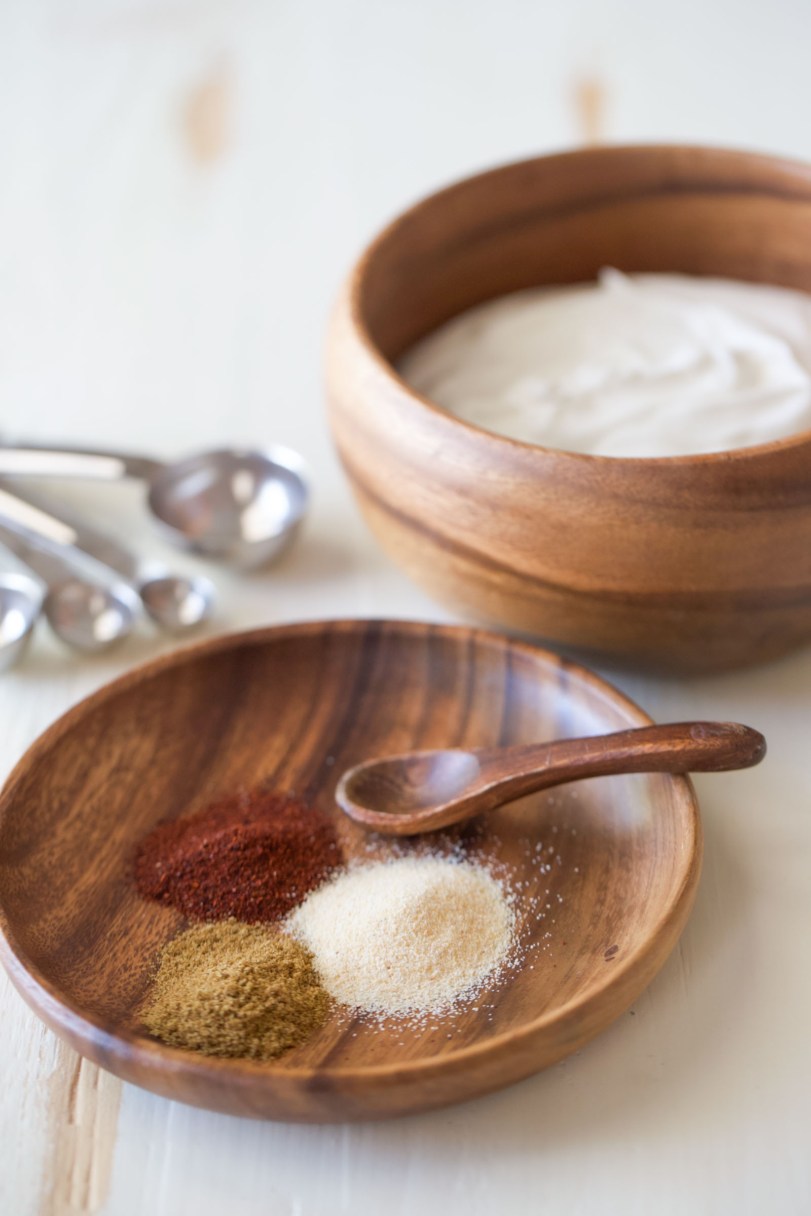 Wooden bowl of sour cream beside a small wooden dish of chili powder, cumin, and garlic powder with a wooden spoon and measuring spoons.