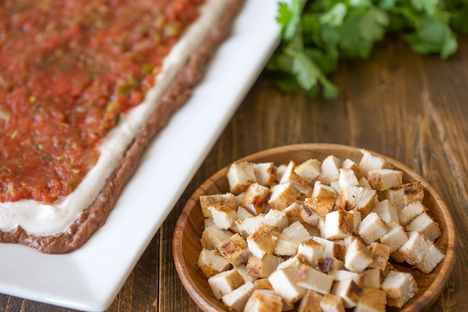 Layers of refried beans, seasoned sour cream, and salsa spread on a white platter next to a wooden bowl of diced grilled chicken.