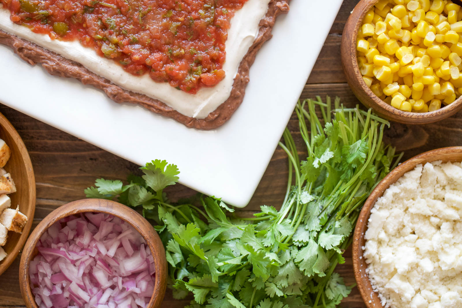 Nine layer dip toppings prepped in wooden bowls — corn, crumbled queso fresco, diced red onion, fresh cilantro, and grilled chicken — on a wood table.