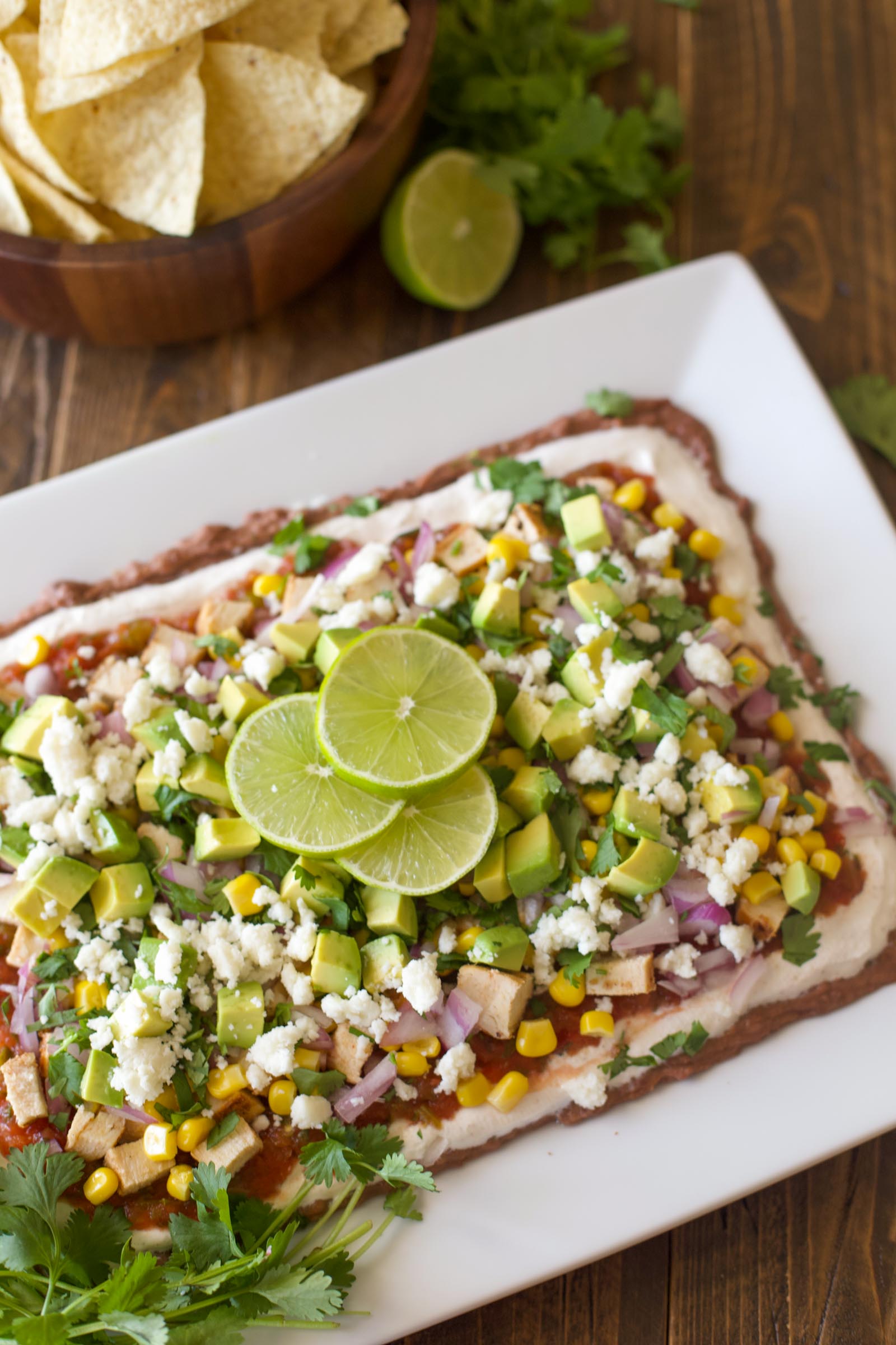 Finished nine layer dip on a white platter garnished with lime slices, served with a bowl of tortilla chips.