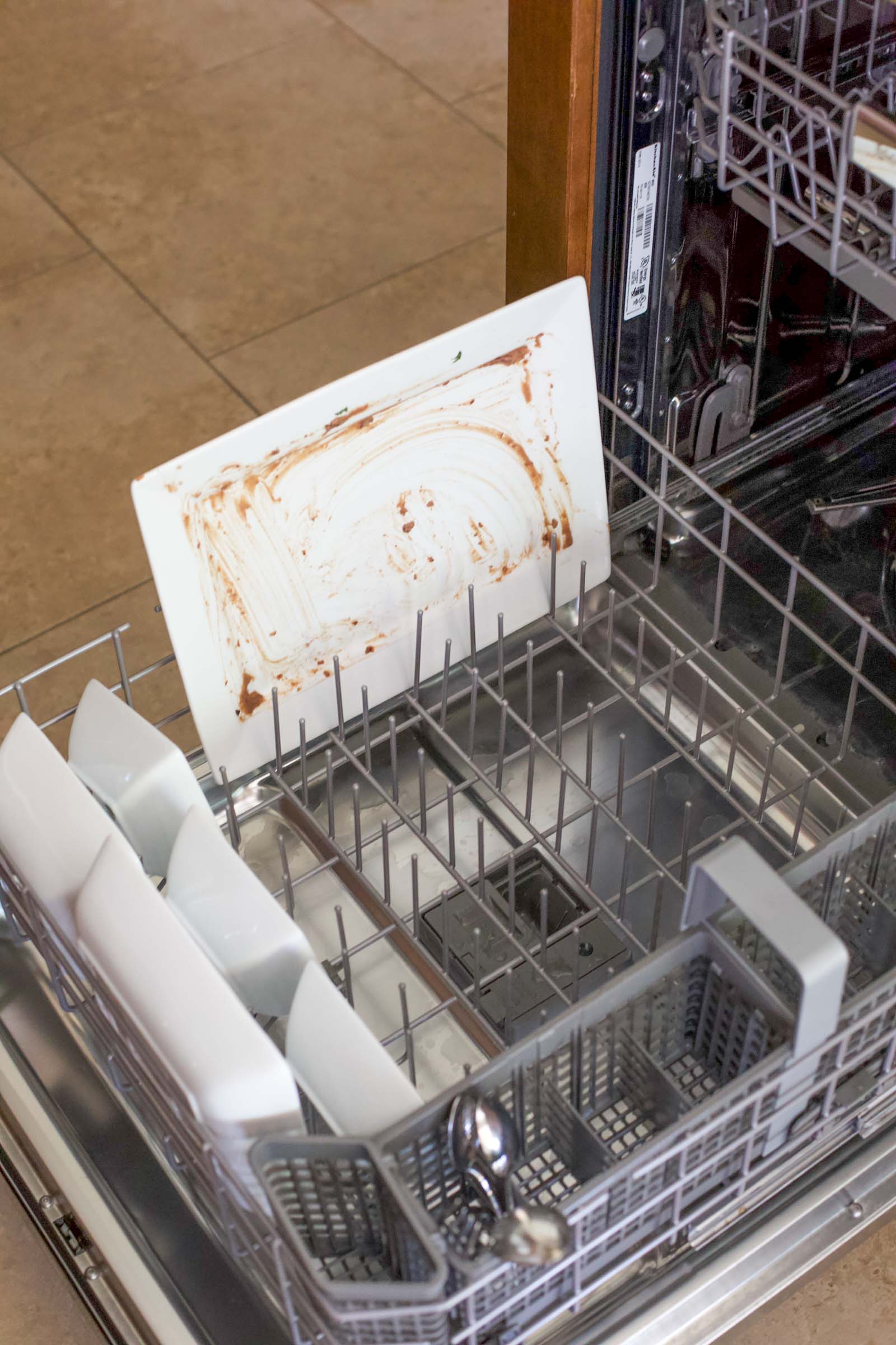 Dirty white serving platter smeared with refried beans loaded into the bottom rack of a dishwasher.